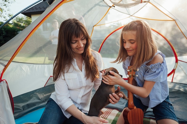 Free photo smiling girl stroking little dog holding by her mother sitting in tent