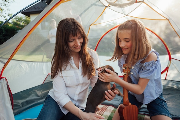 Smiling girl stroking little dog holding by her mother sitting in tent
