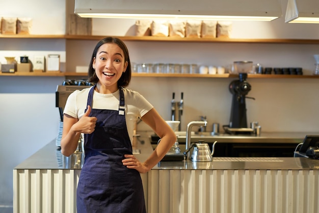 無料写真 カフェバリスタでアルバイトをしている笑顔の女子学生は、コーヒーの近くに立つエプロンを着て親指を立てている