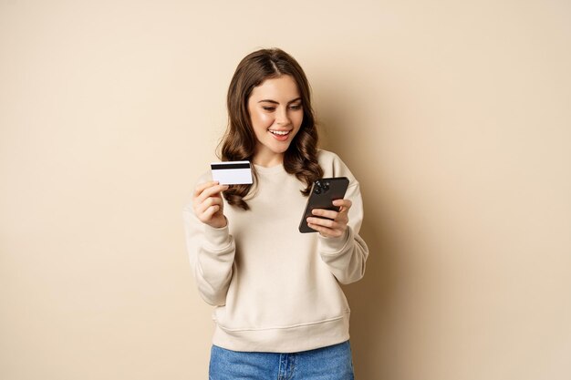 Улыбающаяся девушка с помощью мобильного приложения, покупки смартфона и кредитной карты, стоя на бежевом фоне, заказывает что-то.