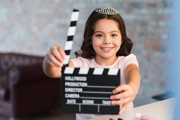 Free photo smiling girl wearing diamond ring crown holding clapper board