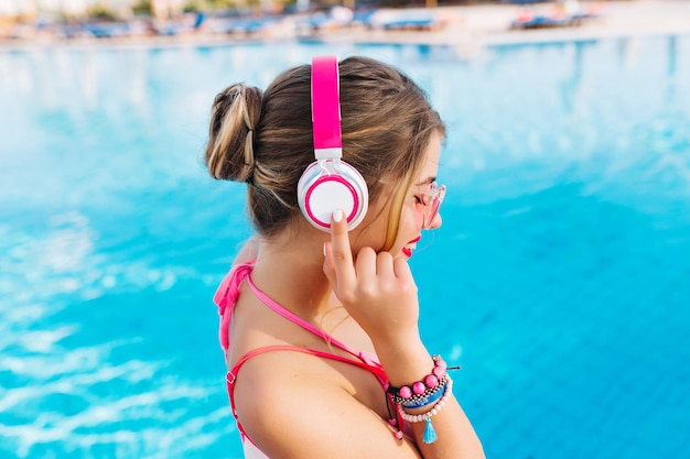 Smiling girl with amazing tan holding new headphones and enjoying weekend on summer resort. Joyful young woman with bright make-up and sunglasses spending time in outdoor pool and listening music