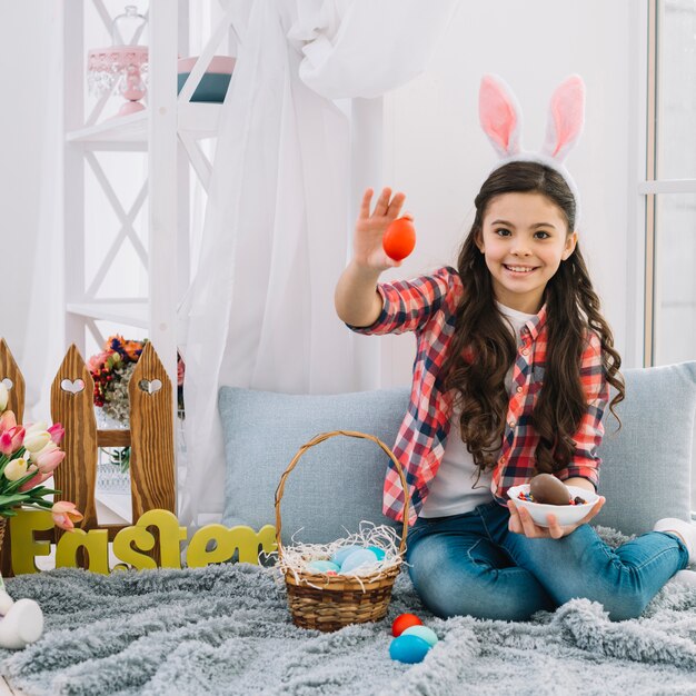 Smiling girl with bunny ears sitting on bed showing red egg on easter day