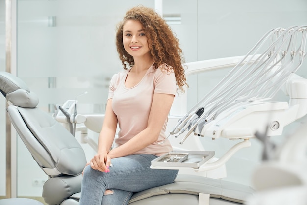 Ragazza sorridente con i capelli ricci che si siede nella poltrona del dentista