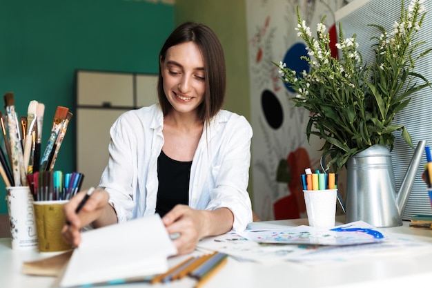 Smiling girl with dark hair sitting at the desk with paintings while happily drawing at cozy home