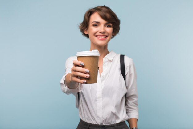 Улыбающаяся девушка с темными короткими волосами в белой рубашке счастливо показывает на камеру чашку кофе, чтобы пройтись по синему изолированному фону