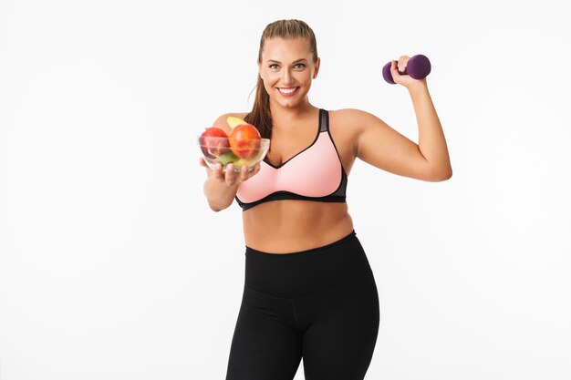 Smiling girl with excess weight in sporty top holding dumbbell in hand while happily showing bowl with fruits on camera over white background