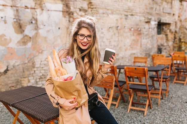 Free photo smiling girl with long curly hair holding bag full of food from market and posing self-consciously. cute young woman  leaned wearily against the fence after shopping. purchase of products, buying meal