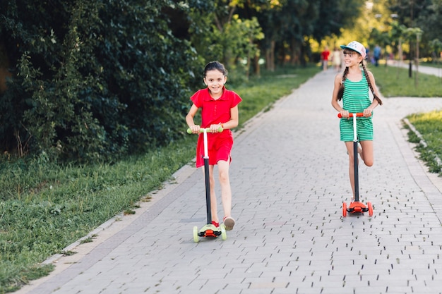 Free photo smiling girls riding on push scooter over walkway in the park