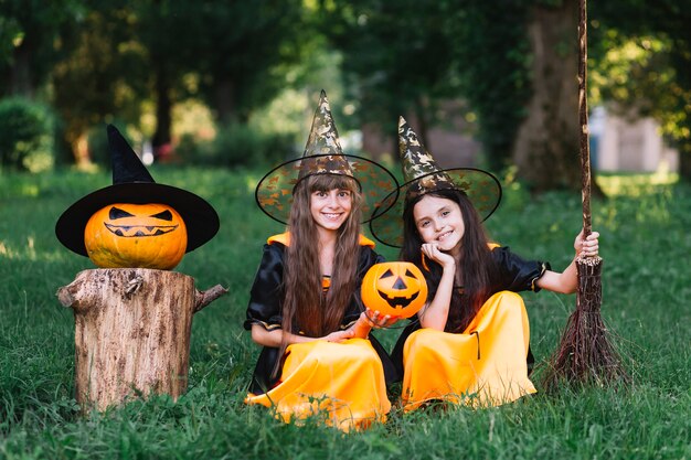 Smiling girls in witch costumes sitting on grass in park 