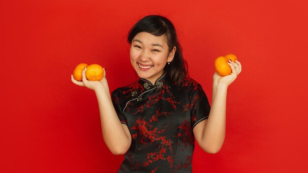 Smiling, giving mandarines. Happy Chinese New Year 2020. Asian young girl's portrait on red background. Female model in traditional clothes looks happy. Celebration, emotions. Copyspace.