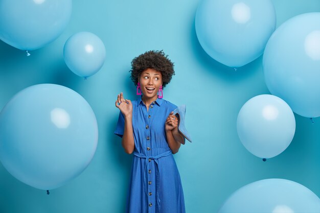 笑顔の嬉しいアフリカ系アメリカ人の女性は、誕生日パーティーの衣装を選び、ドレスに合うようにハイヒールで青い靴を履き、楽しく脇を見て、膨らんだ風船の近くでポーズをとっています。女性、服