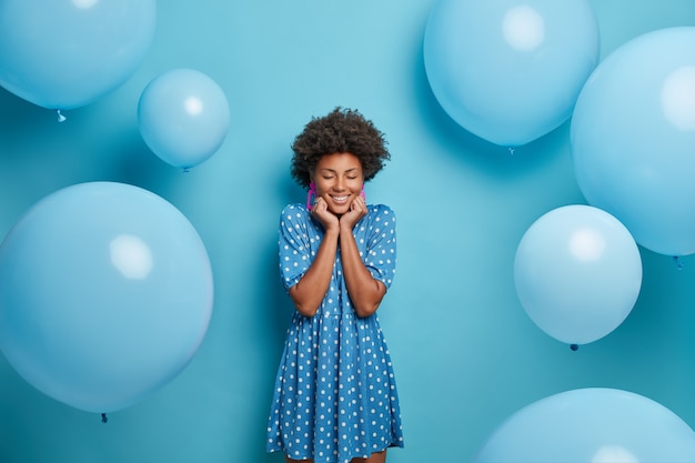 Smiling glad dark skinned woman enjoys birthday party, stands with eyes closed and charming smile, wears fancy blue polka dot summer dress, waits for guests poses around inflated balloons, makes photo