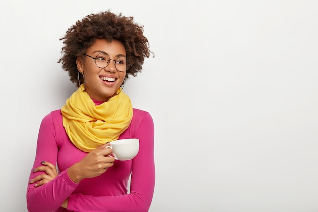 Улыбающаяся счастливая темнокожая женщина держит кружку с ароматным кофе, носит оптические очки, желтый шарф и розовую водолазку, изолированные на белом фоне.