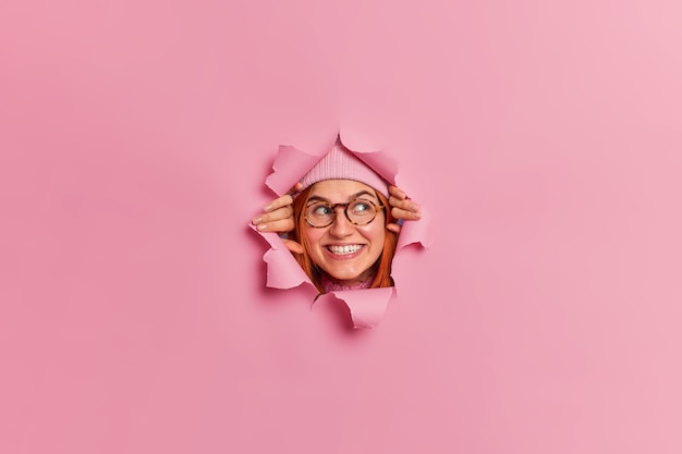 Free photo smiling glad freckled young woman wears optical glasses and looks away through torn paper