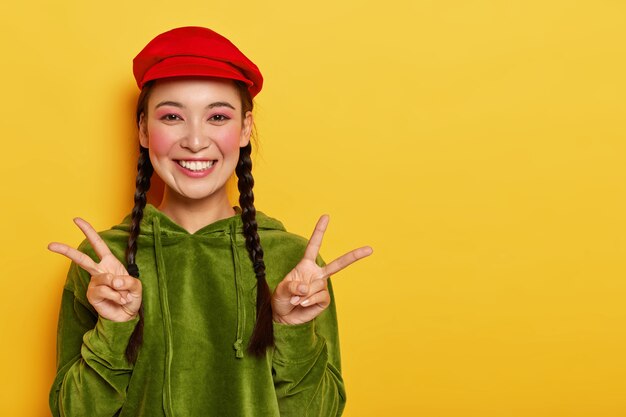 Smiling glad pretty Asian teenage girl shows peace gesture with both hands, smiles broadly