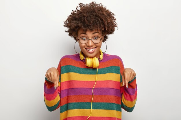 Smiling glad woman points down, proposes cool offer, looks happily at floor, wears transparent eyewear and striped jumper