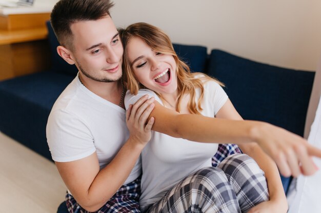 Smiling glad woman in white t-shirt showing something interesting to boyfriend. Indoor photo of refined lady fooling around with husband in morning.