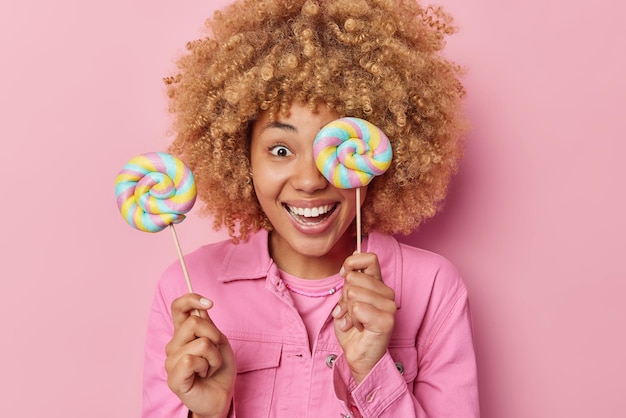 Smiling glad woman with curly hair holds sweet lollipops on sticks enjoys eating favorite treats wears pink jacket poses indoor Positive female model eats caramel candies rewards herself with sweets