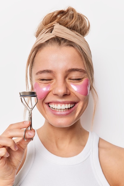 Smiling glad young woman applies hydrogel pink patches takes care of delicate skin around eyes holds eyelashes curler going to do makeup undergoes beauty procedures wears headband and tshirt