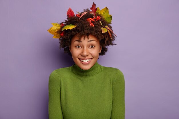 Smiling good looking curly woman being in high spirit, looks happily at camera, wears green turtleneck, autumnal fallen leaves on head, isolated on purple wall
