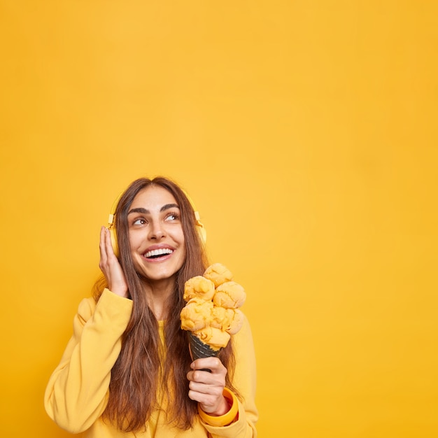 Smiling good looking woman with cheerful expression focused above gladfully holds tasty cone ice cream thinks about something pleasant while listening music via headphones poses against yellow wall