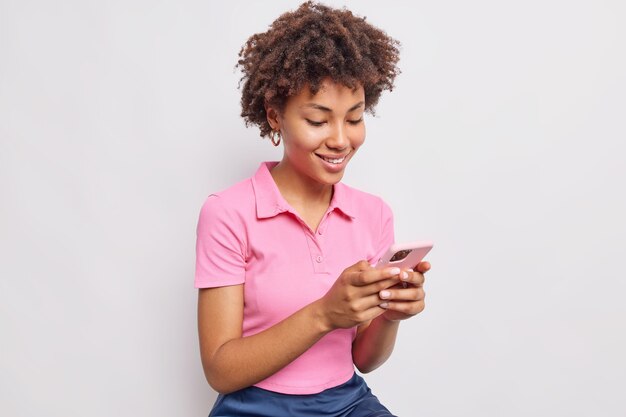 巻き毛の格好良い女性の笑顔は、携帯電話を使用してテキストメッセージを送信し、白い壁に隔離されたカジュアルなピンクのTシャツを着たニュースフィードをスクロールします