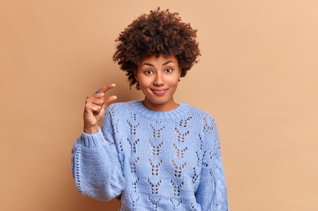 Улыбающаяся симпатичная молодая женщина с вьющимися волосами показывает размерный жест пальцами, формируя что-то маленькое, просит не слишком много носить повседневный свитер, изолированный на коричневой стене студии