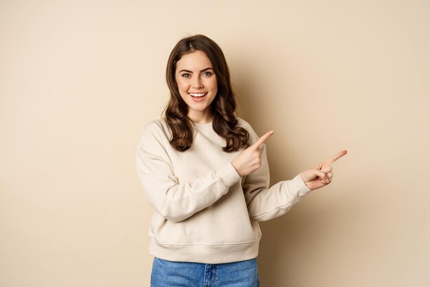 Smiling gorgeous woman pointing fingers right, inviting people, showing logo or banner, standing over beige background