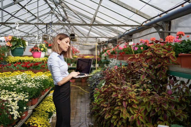 Smiling Greenhouse owner posing with a laptop in her hands talking on the phone having many flowers and glass roof.