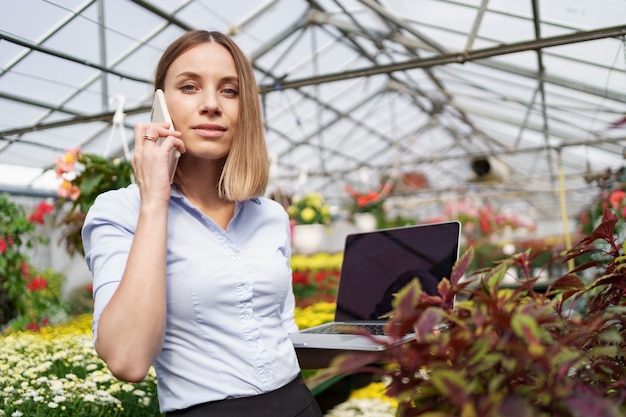 Smiling Greenhouse owner posing with a laptop in her hands talking on the phone having many flowers and glass roof.