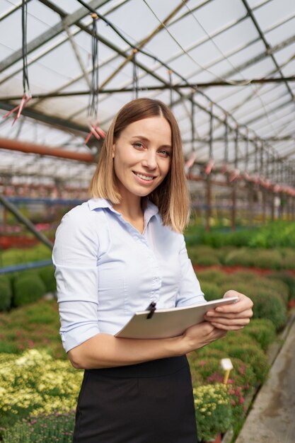 Smiling Greenhouse owner posing with a notepad in her hands having many flowers and glass roof