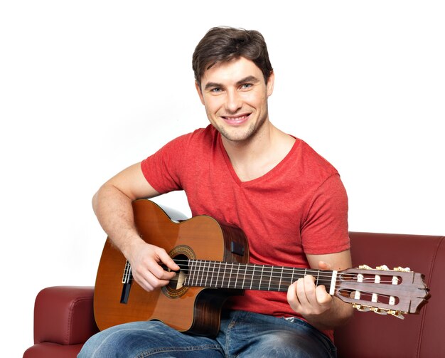 Улыбающийся гитарист играет на изолированной акустической гитаре на белом. Красивый молодой человек сидит с гитарой на диване