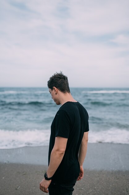 Smiling guy in a black T-shirt stands on the sandy seashore. Summer day, blue sky with white clouds, waves with white foam.