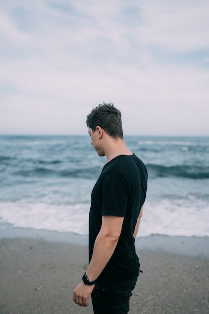 Smiling guy in a black T-shirt stands on the sandy seashore.