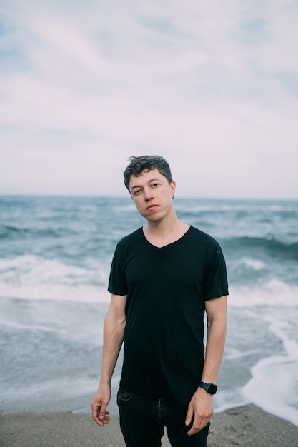 Smiling guy in a black T-shirt stands on the sandy seashore.