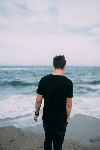 Улыбающийся парень в черной футболке стоит на песчаном берегу моря.