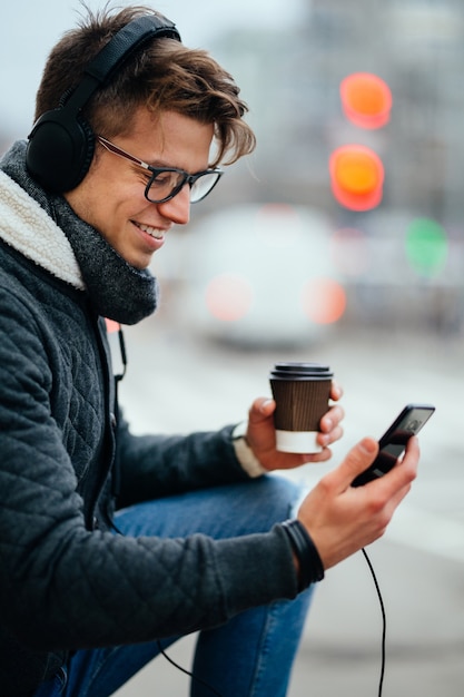 Free photo smiling guy in headphones, using his smartphone, holding a cup of hot coffee, outdoors.