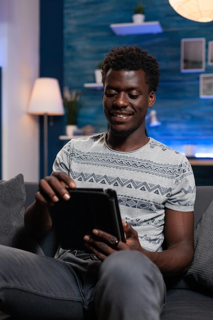 태블릿 컴퓨터를 들고 인터넷에서 검색하는 웃고 있는 남자가 거실에서 집에서 자유 시간을 즐기는 소셜 미디어를 보고 있습니다. 온라인 수업에 대한 정보를 검색하는 프리랜서 남자. 원격 근무