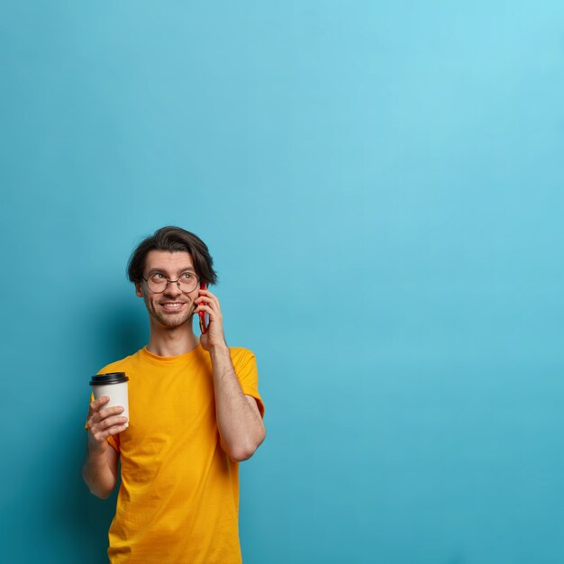 Smiling guy keeps cellular near ear, busy talking with friend, discusses good news, holds takeaway cup of coffee, communicates pleasantly, poses against blue backgroud, copy space for your information