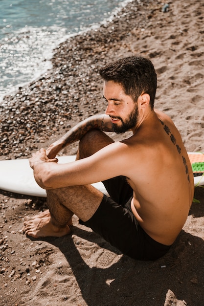 Smiling guy on sandy shore near surfboard