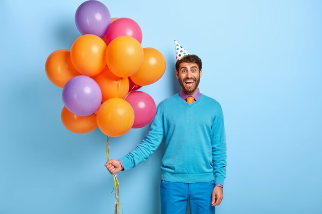 Ragazzo sorridente con cappello di compleanno e palloncini in posa in maglione blu