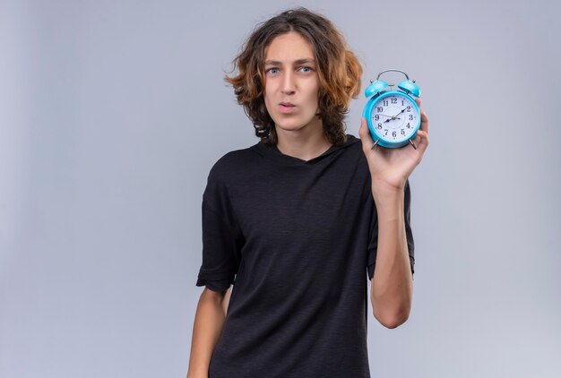 Smiling guy with long hair in black t-shirt holding a alarm clock on white wall