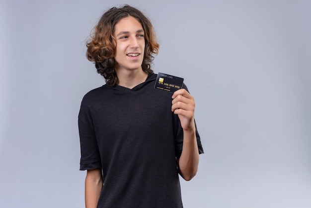 Smiling guy with long hair in black t-shirt holding a bank card on white wall