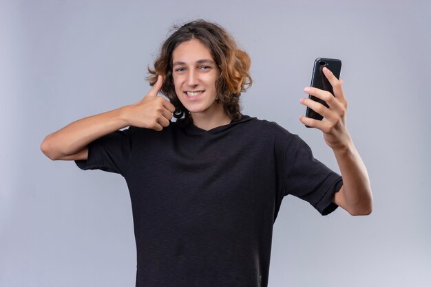 Smiling guy with long hair in black t-shirt holding a phone and shows thumb up on white wall