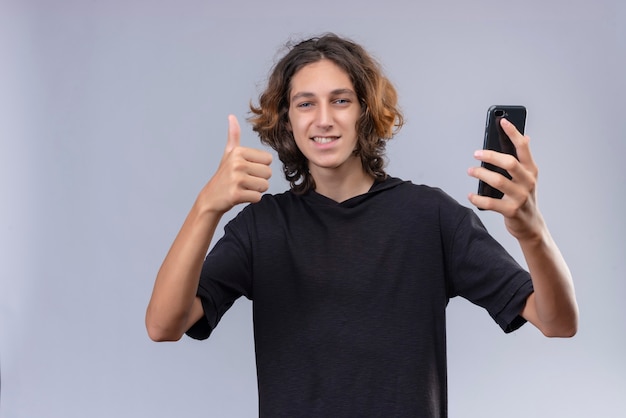 Ragazzo sorridente con capelli lunghi in maglietta nera che tiene un telefono e un pollice in su sul muro bianco