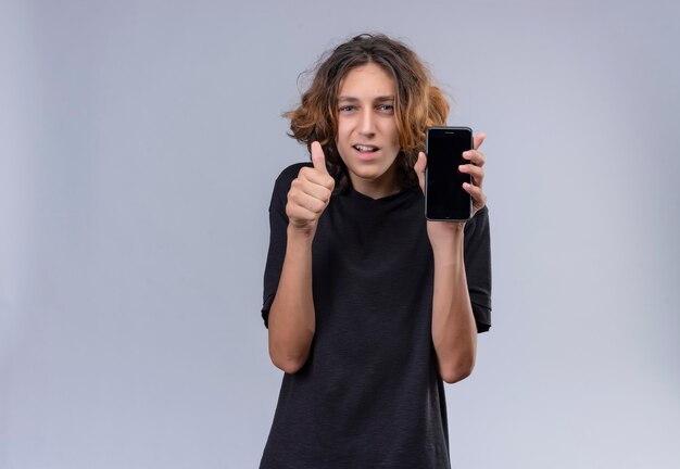 Smiling guy with long hair in black t-shirt holding a phone and thumb up on white wall