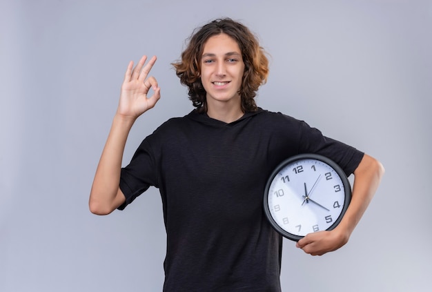 Smiling guy with long hair in black t shirt holding a wall clock and shows okey gesture on white wall