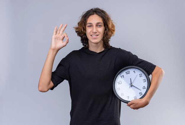 Smiling guy with long hair in black t shirt holding a wall clock and shows okey gesture on white wall