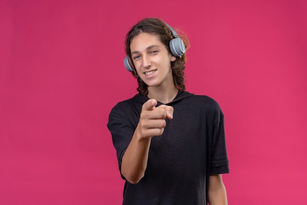 Smiling guy with long hair in black t-shirt listens music from headphone and point to toward ahead on pink wall